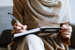 A woman wearing a headscarf writing in a notebook balanced on her lap, to represent CV Writing